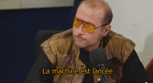 a man wearing sunglasses and a leather vest is sitting in front of a microphone and the words la machine est lancée are above him