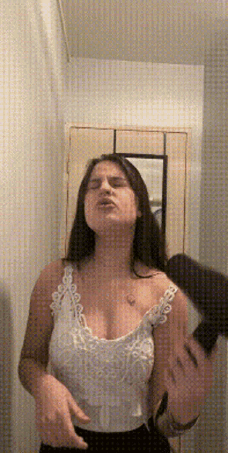 a woman in a white tank top is standing in front of a mirror with her eyes closed .