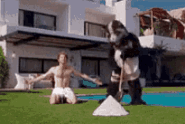 a shirtless man is kneeling on the grass in front of a house while a dog stands behind him