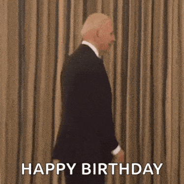 a man in a suit and tie is standing in front of a curtain and saying happy birthday .