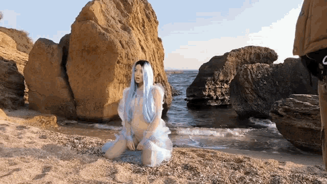 a woman in a blue dress sits on the beach near a large rock
