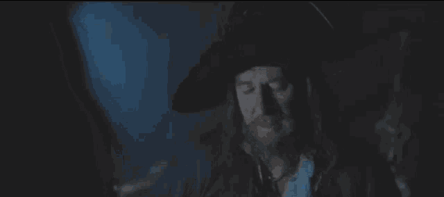 a man with long hair and a beard is wearing a hat and a necklace in a dark room .