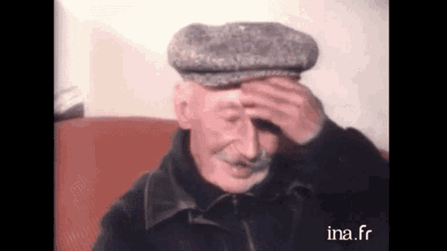 an old man wearing a hat is sitting on a couch and covering his face .