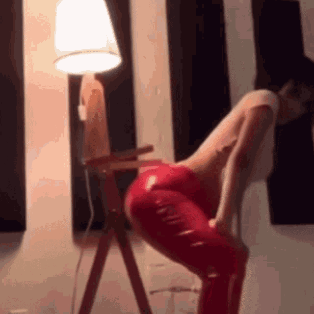 a woman in red leather pants is squatting down in front of a lamp in a room .