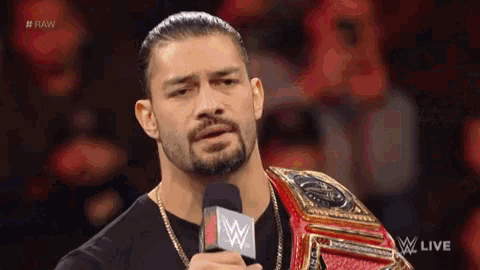roman reigns is holding a microphone while wearing a wrestling championship belt .