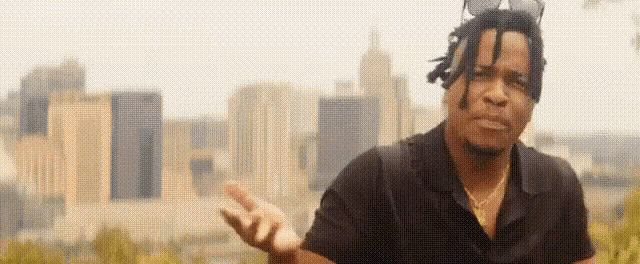 a man with dreadlocks is standing in front of a city skyline and making a peace sign .