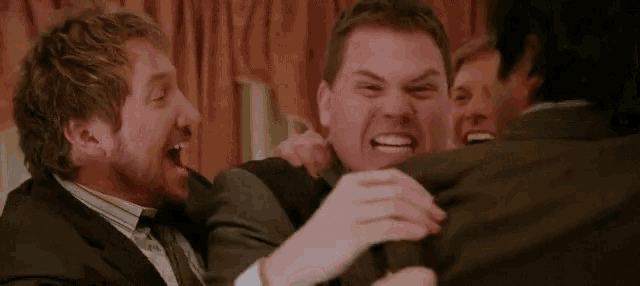 a group of men are laughing and hugging each other . one of the men is making a funny face .