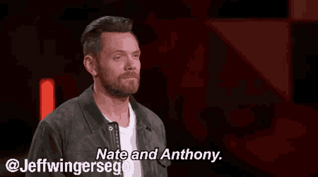 a man with a beard and a leather jacket is standing in front of a red wall and talking to someone .
