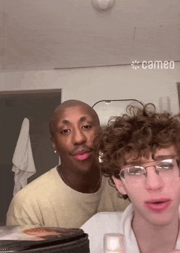 a man with curly hair is standing next to another man with glasses and a cameo logo on the wall behind them