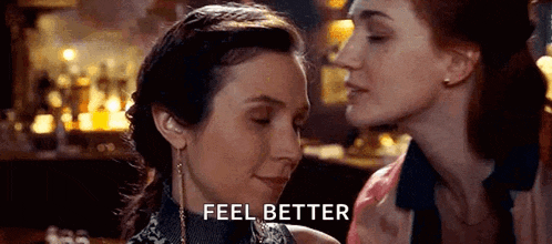 a woman is kissing another woman on the cheek in a bar while another woman says `` feel better '' .