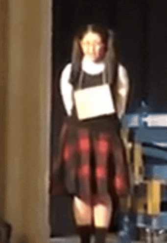 a girl in a plaid skirt is standing on a stage holding a piece of paper