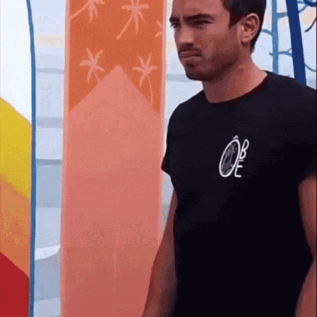 a man wearing a black oe shirt stands in front of a surfboard