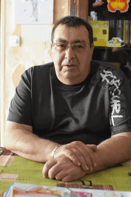 a man sits at a table with his hands folded wearing a black shirt with chinese writing on the sleeves