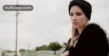 a woman wearing a black hat and a fur coat is standing in front of a pole and says `` stupid bitch '' .