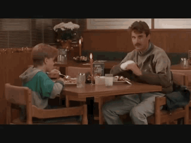 a man and a boy are sitting at a table in a restaurant with a box of cornflakes on the table