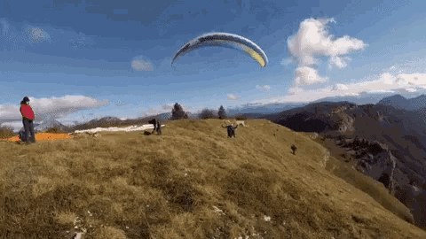 a paraglider flies over a grassy hillside with mountains in the background
