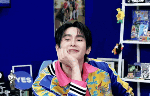 a young man in a colorful jacket is sitting in front of a shelf with his hand on his chin .