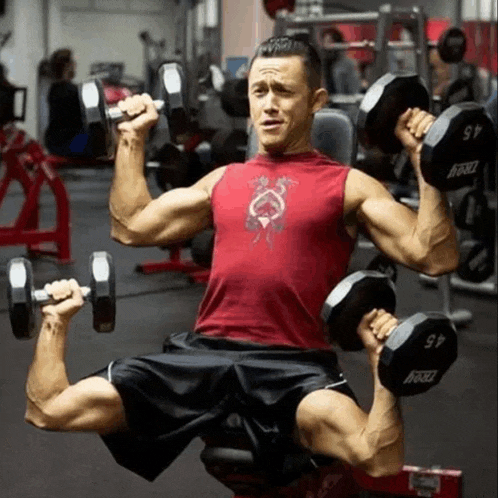 a man in a red tank top is lifting dumbbells in a gym ..