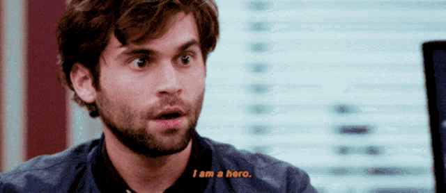 a man with a beard says " i am a hero " in front of a window