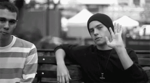 a black and white photo of two men sitting on a bench . one of the men is wearing a beanie .