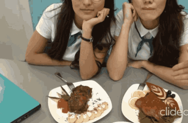 two girls are sitting at a table with plates of food and the word clideo on the bottom left