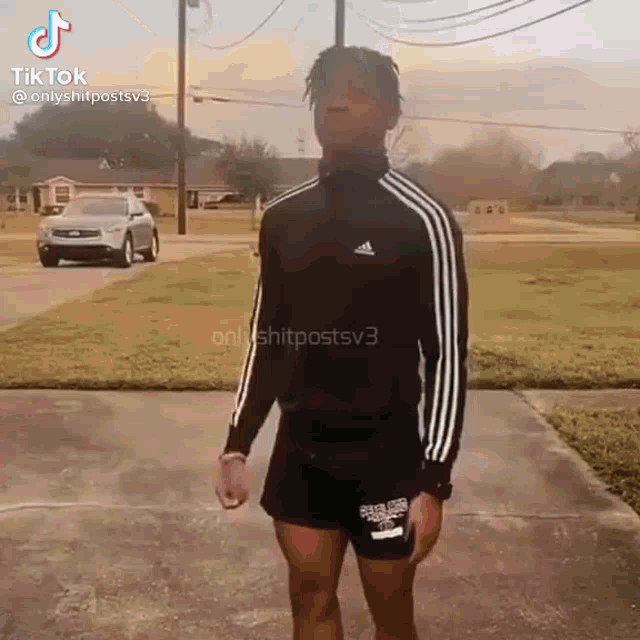 a man in a black adidas jacket and shorts is standing on a sidewalk in front of a car .