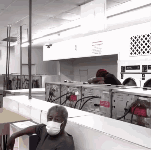 a man wearing a mask sits in a laundromat with a sign on the wall that says no smoking