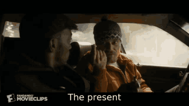 two men are sitting in a car with the present written on the bottom