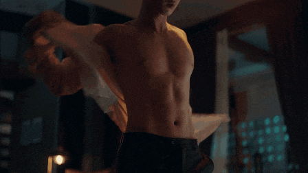 a shirtless man stands in a dark room with his sleeves rolled up