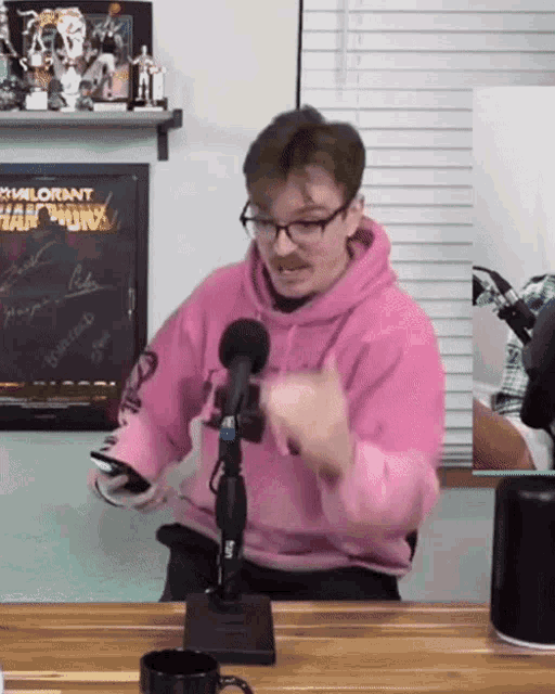 a man wearing a pink hoodie and glasses is sitting in front of a microphone .
