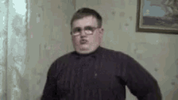 a man wearing glasses and a sweater is standing in front of a painting .
