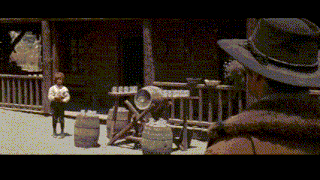 a man in a cowboy hat is standing in front of a building with barrels and a boy standing on the porch .