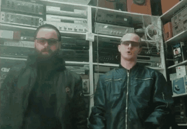 two men with beards and sunglasses are standing next to each other in a room filled with lots of electronics .