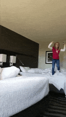 a little girl in a red shirt jumps in the air in a hotel room