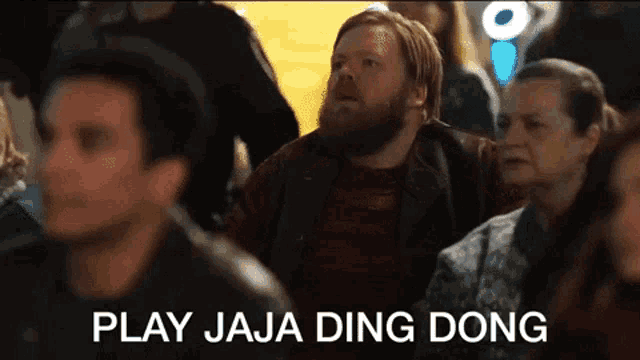 a man with a beard is sitting in a crowd with the words play jaja ding dong written on the screen .