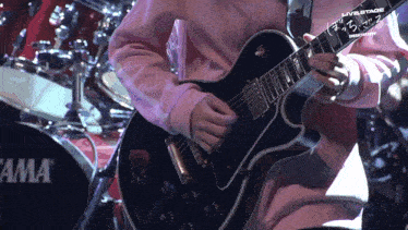 a person in a pink sweatshirt is playing a guitar in front of a tama drum set