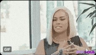 a woman with blonde hair is sitting in front of a window with her hands on her chest .