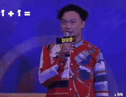 a man in a red shirt is holding a microphone in front of a blue background that says 1 + 1 = ?