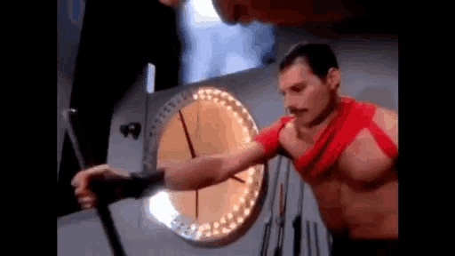 a man without a shirt is standing in front of a clock and holding a shield .