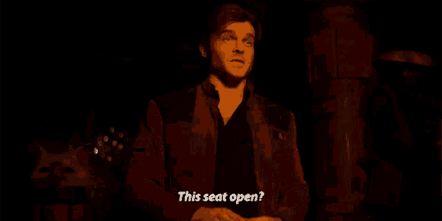 a man in a red jacket is standing in a dark room and says this seat open .
