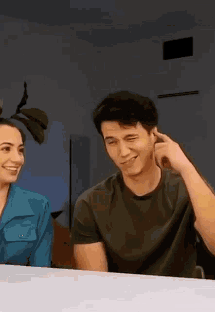 a man and a woman are sitting at a table and smiling . the man is touching his ear .