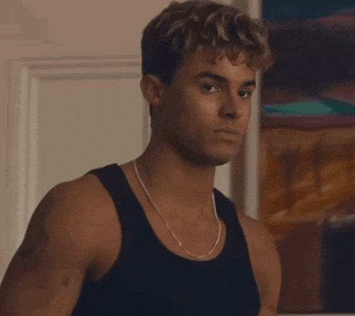 a young man wearing a black tank top and a chain around his neck looks at the camera