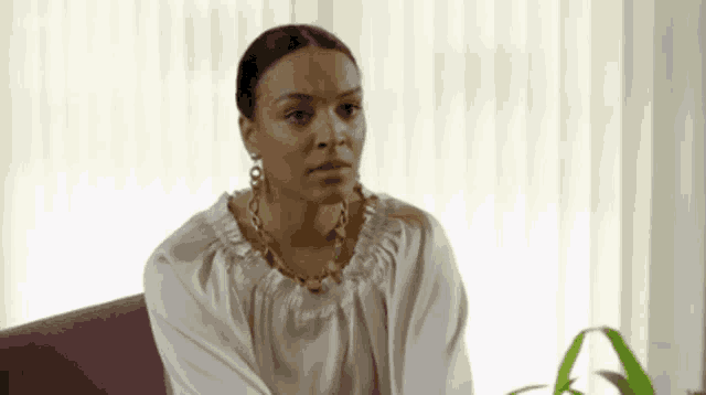 a woman is sitting on a couch in front of a window wearing a white shirt and earrings .