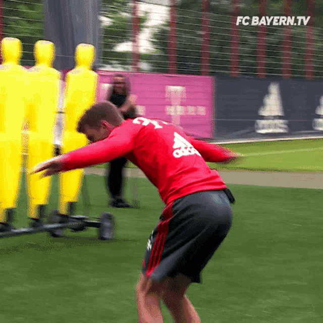 a man in a red shirt and black shorts is doing exercises on a soccer field with yellow mannequins .