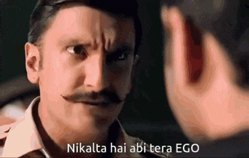 a man with a mustache is talking to another man while making a funny face .