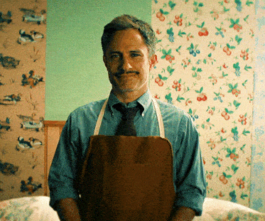 a man wearing an apron and tie stands in front of a wall with floral wallpaper
