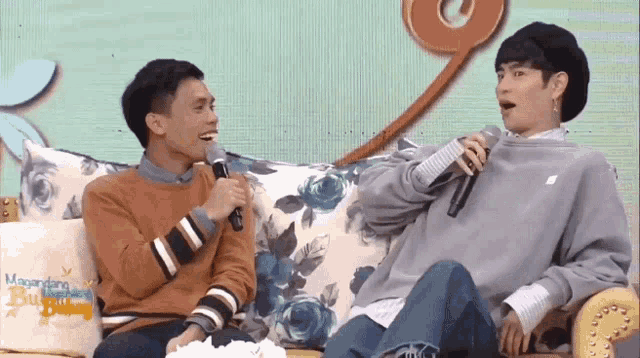 two men are sitting on a couch talking to each other with microphones in their hands .