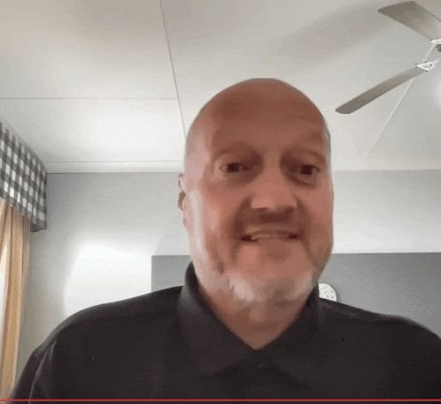 a bald man with a beard wearing a black shirt is smiling in front of a ceiling fan