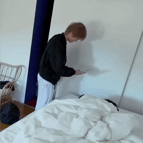 a man in a black jacket is standing next to a bed looking at his phone
