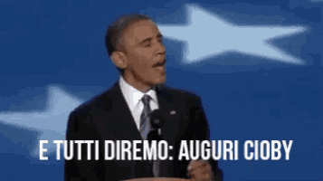 barack obama is giving a speech in front of a flag and says e tutti diremo